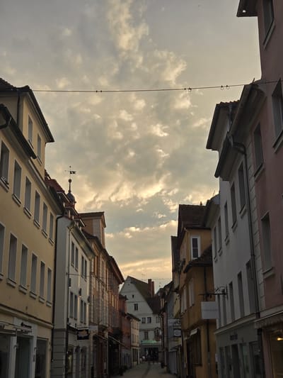 The photograph captures a street scene in Ansbach, Germany, likely during the late afternoon or early evening, given the soft, diffused light and the dramatic, cloudy sky. The perspective is looking down a narrow street lined with historic buildings on both sides. The architecture suggests an older European town, with multi-story buildings featuring pitched roofs, prominent window frames, and varied facades in muted colors like yellow, cream, white, and pink.
On the left side of the street, a pale yellow building dominates the foreground, with several tall, rectangular windows visible. Adjacent to it, a white building with grey detailing follows.
On the right side, a pink-colored building begins the row, followed by a white building, and then a yellow building with a sign that reads "horand.foto." Further down the street, other buildings are visible, with signs and shopfronts at ground level, indicating commercial activity.
A prominent element in the sky is a wire strung across the street, with what appear to be insulators or rings attached. The sky itself is filled with textured, grey and yellowish clouds, suggesting the presence of the sun behind them. On the roof of one of the buildings in the mid-ground, a weather vane is visible.
In the distance, at the end of the street, a few figures are walking, suggesting the presence of people, though they are too indistinct to identify. The overall atmosphere is one of quiet, historic charm, with a touch of atmospheric drama provided by the sky. The street appears to be paved, and the scene is framed by the buildings, creating a sense of depth and perspective.