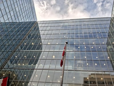 The image shows a modern glass-facade building, likely an office building, in Ottawa, Canada. The building has a geometric design with many glass panels and visible interior lights reflecting on the exterior. In the foreground, a Canadian flag is visible on a flagpole, partially obscuring the building's reflection. The sky above is cloudy, with a mix of grey and white clouds. The perspective is from a low angle looking up at the structure.