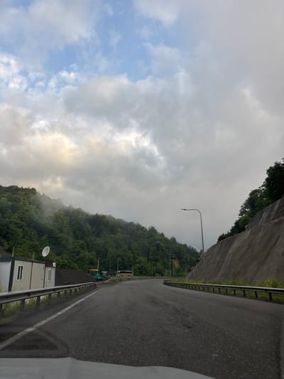 The image captures a curved asphalt road winding through a mountainous, forested landscape under a cloudy sky. On the left side of the road, there is a white modular building with a satellite dish on its roof. Behind the building, a pile of dark gravel and construction equipment, including a yellow asphalt roller and a green excavator, are visible. Further down the road on the left, a yellow bus is parked. The right side of the road is bordered by a steep, grey retaining wall with trees and vegetation growing on top. A lone street lamp stands along the right edge of the road. The scene appears to be in a rural or semi-rural area, possibly undergoing construction or maintenance. The location is identified as Unknown, Georgia.