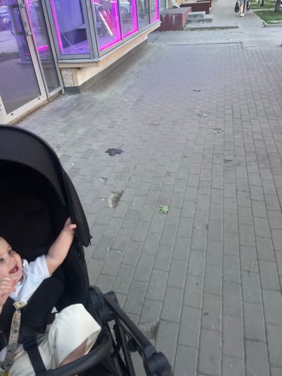 A baby is in a stroller on a sidewalk in Novopodkletnoye, Russia. The baby is wearing a white t-shirt and a white hat, and is smiling and reaching out with their right hand. The stroller is black. The sidewalk is paved with gray rectangular bricks. There are some leaves on the sidewalk. To the left of the sidewalk is a building with large windows that have pink neon lights. The building appears to be a commercial establishment. In the background, there are trees and another person walking on the sidewalk. The lighting suggests it is late afternoon or early evening.