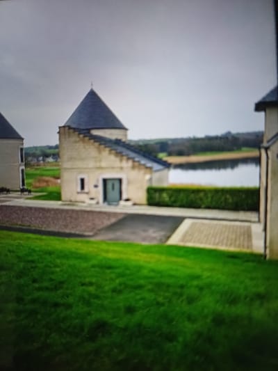 The image depicts an exterior scene on an overcast day, likely near Enniskillen, United Kingdom, specifically along Lough Erne. In the midground, a distinctive light-colored building stands centrally, featuring a dark grey conical roof topped with a finial and a prominent dark grey stepped gable or parapet descending from the roofline on its left side. This building has a dark grey entrance door and at least one small rectangular window. To its left, a dark gravel area is visible, while a dark asphalt driveway and an adjacent light-colored block-paved section lead to the building's entrance. A manicured dark green hedge runs horizontally behind the building's property line.
To the immediate left and right, parts of similar light-colored buildings with dark roofs are partially visible, framing the central structure. The foreground consists of a vibrant green lawn sloping upwards towards the viewer. Behind the hedge in the background, a calm body of water (Lough Erne) stretches, reflecting the grey, overcast sky. On the far bank of the water, a distant treeline, green fields, and faint structures are discernible under the uniform grey sky. No people or vehicles are present in the scene.