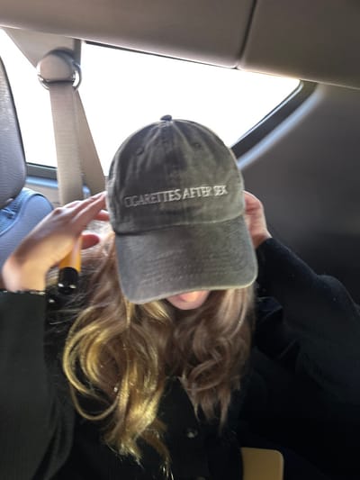 Inside a vehicle, a person wears a gray baseball cap with "CIGARETTES AFTER SEX" embroidered on the side. Light-brown hair flows beneath the cap. The person wears a black jacket or coat and holds a yellow object in their hand. A seatbelt is visible, and sunlight streams through the car window. The image has been rotated counter-clockwise.