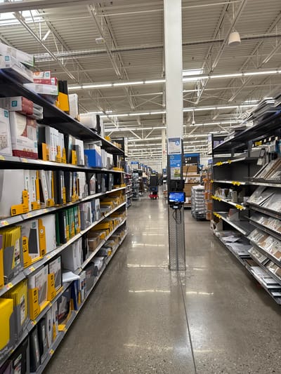 This image captures a long, brightly lit aisle within a large retail store, identified as a Walmart, in Worcester, United States. The perspective looks down the length of the aisle, with shelves of merchandise lining both the left and right sides, receding into the distance.

The store's ceiling is high, featuring exposed white structural beams, trusses, and numerous fluorescent lighting fixtures that provide ample illumination, which is reflected on the polished, light-colored concrete floor.

The shelves are well-stocked with what appear to be office supplies, including binders, folders, and various paper products. Many items are packaged in white, black, blue, and yellow, with some featuring yellow information or price tags. Visible text on product packaging includes "DURABLE VIEW BINDER," "DURABLE VIEW," "MAO," and "ESSENTIALS."

Mid-way down the aisle, a blue sign bearing the "Walmart" logo is suspended from the ceiling. This sign also displays text indicating "Office Supplies" (partially visible) and an aisle number, possibly "Aisle 37" or "Aisle 7." Below this sign, a digital display or kiosk is mounted on a tall, dark pole, likely for customer information or self-checkout.

No people are visible in the aisle, creating a sense of quiet and order. The bright artificial lighting suggests the store is open or ready for business, though the absence of shoppers could indicate an off-peak time, such as early morning or late evening. The scene is purely functional, devoid of any discernible activity or emotion.