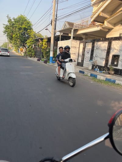 Two individuals are riding a white scooter down a paved street in Sugih Waras, Indonesia. The person in front is wearing a black hoodie and jeans, while the person behind them is wearing a blue shirt with white writing and dark shorts. Both are wearing helmets. The scooter has a white body and silver accents. The street is lined with houses on the right side of the image, some of which have modern architectural elements and decorative marble facades. There are trees and utility poles along the street. The sky is clear and a light blue, indicating it is daytime. The image appears to be taken from another moving vehicle, as a portion of a side mirror is visible in the lower right corner.