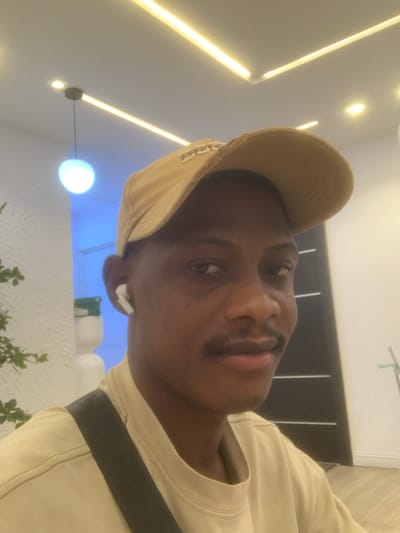 The image captures a close-up, angled portrait of a young man, presumably indoors. The man has a dark complexion and is looking directly at the camera with a neutral to slightly smiling expression. He is wearing a light beige or tan baseball cap, a light-colored (possibly cream or beige) t-shirt, and a dark shoulder bag strap diagonally across his chest. A white wireless earbud (AirPod) is visible in his right ear. He has a mustache and a faint goatee.
The background shows a modern interior setting. The ceiling features warm-toned linear LED strip lights and a suspended spherical light emitting a soft glow. Further in the background, a distinct blue ambient light emanates, possibly from an accent light or a light-colored object illuminated by blue light. Below the blue light, there's a white, textured surface, possibly a wall or a decorative planter, adorned with some green foliage. A dark, horizontal structure, perhaps a partition or a piece of furniture, is visible behind the man's head and shoulder. The overall lighting suggests an artificial indoor environment. There is no visible text in the image. The image is presented at an angle, making it appear as if the man's head is tilted to the right. The activity appears to be posing for a photograph. The location context is Animashawun, Nigeria.