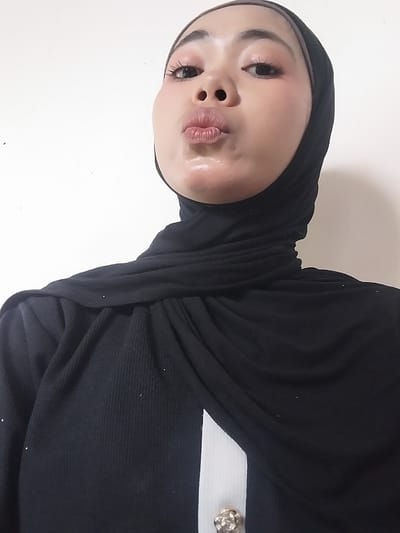A close-up, head-and-shoulders shot captures a young Indonesian woman, adorned in a black hijab and a black ribbed top with a white stripe down the center. Her face is turned towards the camera, with her lips pursed in a kissing gesture, glistening with a clear lip gloss. Her eyes are open, and she has makeup on, including eyeliner and blush. She appears to be indoors, against a plain white background, with no discernible city features from Jeppee, Indonesia, visible in the image.