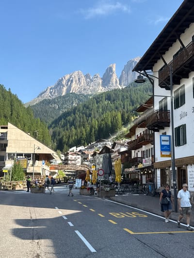 The image depicts a street scene in Campitello di Fassa, Italy, with a prominent view of the Dolomite mountains in the background. The foreground shows a paved road with yellow and white road markings, including a "BUS" designation painted on the asphalt. Several pedestrians are visible walking on the sidewalk and crossing the street. Buildings with traditional alpine architecture, featuring balconies and wooden accents, line the street. Outdoor seating for cafes or restaurants is set up along the sidewalk. The background is dominated by a majestic mountain range covered in dense evergreen forest, under a clear blue sky. The sun is casting shadows on the road, indicating it is daytime. A bus stop sign is mounted on a pole.