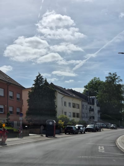 The image captures a detailed street scene in Sanem, Luxembourg, under a bright, partly cloudy sky. The perspective is from the road, looking slightly uphill as the asphalt road curves gently to the right. The sky in the upper portion of the frame features a light blue color with scattered white cumulus clouds. Multiple faint and distinct white contrails from aircraft are visible, crisscrossing the sky. On the left side of the road, in the foreground, a red and white construction barrier with an amber warning light is present. Behind it, a yellow fire hydrant or utility cap is visible. Further back, a dark grey portable toilet with a red door and a white label is situated on a paved area. Adjacent to it, a pole supports multiple traffic signs, including a blue circular sign with a red diagonal line, indicating a no-stopping or no-parking zone. The sidewalk area appears clean and paved. The closest building on the left is a reddish-brown, multi-story residential structure with a dark tiled roof. Its facade features multiple rectangular windows fitted with white frames and white external roller shutters, some partially open. A small, manicured green shrub is visible in front of this building. A large, dark green coniferous tree stands between this building and a lighter-colored building further back, partially obscuring the latter. The dark grey asphalt road occupies the central and lower portions of the image, showing a slight sheen. White road markings, including diagonal lines and an inverted "STOP" legend, are visible on the right side of the road. A grey curb separates the road from the adjacent paved areas. On the right side of the road, a series of multi-story residential buildings line the street. These buildings display facades in varying shades, including light cream, light grey, and white. They feature dark tiled roofs, some with dormer windows and balconies with dark railings on the grey building. Several cars are parked parallel to the curb along this side of the road. From foreground to background, visible vehicles include a dark grey Volkswagen Golf (Luxembourg license plate MB 7282), a white SUV (Luxembourg license plate LU 0725), a dark grey SUV (Luxembourg license plate GD 9100), and additional less distinct cars further along the curve. A dark sedan is visible at the far end of the road curve. Behind the buildings and cars on the right, lush green deciduous trees and