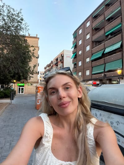 A young woman with blonde hair is taking a selfie in an urban environment. She is wearing a white, eyelet-detailed top. Her expression is a slight smile. She is positioned in the foreground, with a street and buildings in the background. To her left, there is a tree, a lamppost, a green awning over a shop with a sign that appears to say "Iberdrola", and a grey trash bin. To her right, there is a large, grey dumpster with some text and images visible on its side. Further in the background, there are multi-story brick buildings with balconies and green awnings. The sky above is a clear, pale blue, suggesting it is either early morning or late afternoon. The scene appears to be in the Benimàmet-beniferri district of Valencia, Spain, given the presence of the "Iberdrola" sign, a Spanish energy company.