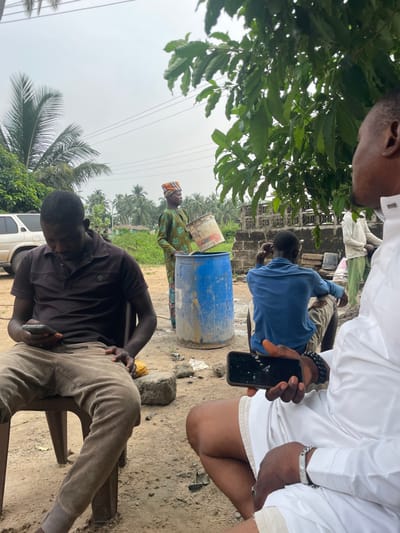 The image depicts an outdoor scene in Ganyingbo, Nigeria, under an overcast sky. The foreground shows an unpaved, light brown dirt ground with scattered debris.
Five adult males are present. In the immediate foreground left, a dark-skinned man wearing a dark brown polo shirt and light brown pants sits on a brown plastic chair, looking down at a dark-colored smartphone held in both hands. To his right, a partial view of another dark-skinned man shows him seated, wearing a white robe-like garment and white shorts. He holds a black smartphone with a cracked screen in his left hand and wears a black beaded bracelet on his left wrist and a silver-colored chain bracelet on his right wrist.
In the midground, a dark-skinned man in a patterned green long-sleeved garment and an orange and brown headwrap stands, facing right. He holds a white plastic bucket tilted over an upright blue plastic barrel, appearing to pour contents into it. Behind the barrel, another dark-skinned man in a blue long-sleeved shirt and light-colored pants is seated on a low object, facing away from the viewer. Further in the background right, partially obscured by green foliage and a concrete block wall, a fifth dark-skinned man in a light-colored long-sleeved shirt stands.
The background features lush green vegetation, including numerous palm trees and other broad-leafed trees. A white SUV is partially visible in the far left background. Overhead power lines traverse the grey, cloudy sky.