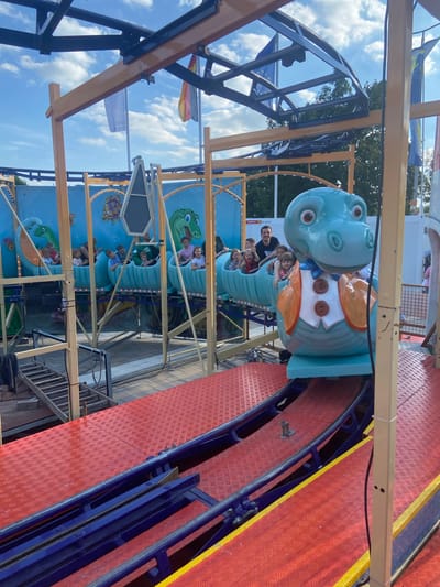 The image depicts a brightly colored amusement park ride in Baunatal, Germany, likely at the Loxx Miniatur Welten attraction, given the German flags visible. A blue, dinosaur-themed roller coaster car, designed with large cartoon eyes and wearing a vest and bow tie, is the primary subject. Several children, appearing to be between the ages of five and ten, are seated in the front cars of the roller coaster, some smiling and looking towards the camera. A man is visible in the background, also seated in one of the cars. The roller coaster track is a vibrant red with blue railings. The ride is supported by a wooden and metal structure, and a blue backdrop with cartoon underwater scenes, featuring more dinosaur characters and sea creatures, adorns the rear of the ride. The sky above is partly cloudy.
