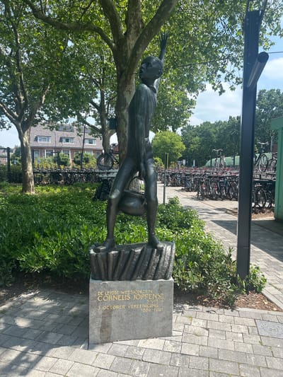 A bronze statue of a man stands on a stone pedestal in front of lush green bushes. The statue depicts a lean figure with one arm raised and the other holding a pot. The pedestal is inscribed with gold lettering that reads, "DE LEIDSE WEESJONGEN CORNELIS JOPPENSZ 3 OCTOBER 1574 3 OCTOBER VEREENING 1886-1931". Behind the statue, a large number of bicycles are parked in racks, with a building and trees visible in the background. The scene appears to be in an urban area, possibly a public square or park in Leiden, Netherlands. The ground is paved with grey bricks.
