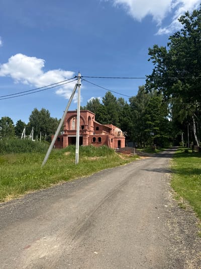 The image depicts a rural road leading towards a partially constructed brick building. The building, made of red brick, features arched windows and doorways and appears to be in the early stages of construction. Several power lines are visible, supported by a concrete pole in the foreground. The road is unpaved, with gravel and dirt. Surrounding the road and building are lush green grass and trees, suggesting a natural, undeveloped environment. The sky is blue with scattered white clouds. The scene is static, with no people or animals visible. The location is identified as Гаранскі сельскі Савет, Belarus.