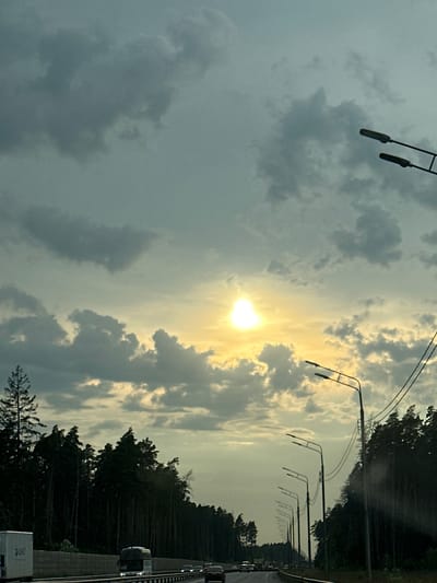 This is a photograph taken from a vehicle on a highway, likely in or near Khliupino, Russia. The scene depicts a cloudy sky with the sun peeking through, casting a bright glow. A dense forest of pine trees lines the right side of the highway, with a few trees visible on the left. Several streetlights are positioned along the road, and power lines are strung between them. The highway is a multi-lane road with traffic moving in the same direction. Several vehicles, including trucks and cars, are visible on the road. The overall atmosphere is somewhat overcast, with the sun providing a dramatic contrast against the dark clouds and trees.