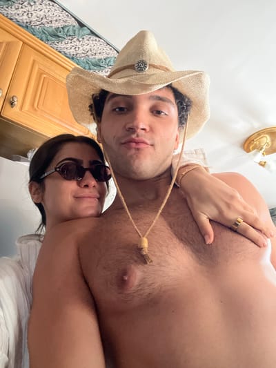 A selfie taken indoors. A light-skinned man wears a straw cowboy hat. A light-skinned woman wears sunglasses. The man's chest is exposed. A woman's hand with a gold ring rests on his chest. The room appears to have a wooden cabinet and a light fixture. There's no clear indication of a location in Èze, France.