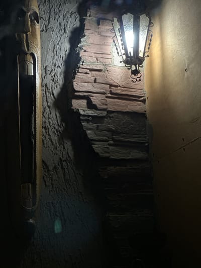 The image shows a dimly lit, narrow passageway with textured walls. On the left, a carved wooden mask is mounted vertically, its features partially obscured by shadow. Adjacent to the mask is a rough, grey, textured wall. To the right of this wall is a section of stacked stone, appearing to be a rough-hewn reddish-brown stone. Above this stone wall, an antique lantern with ornate metalwork and glass panels illuminates the scene. The light from the lantern casts a strong shadow on the textured wall. To the far right, a smooth, pale yellow wall is visible, with a faint shadow extending downwards from the lantern. The overall impression is one of an interior, possibly a historic building or themed environment. No people or discernible actions are present. There is no information in the image or its crops to indicate the location is Slov'yanoserbs'k, Ukraine.