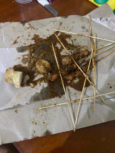 The image shows a messy pile of chicken satay on brown paper, likely in an outdoor or casual setting in Karimunjawa, Indonesia. The satay skewers are scattered haphazardly, with some pieces of chicken and sauce spilled on the paper. There are also some pieces of what appear to be lontong (rice cakes) among the satay. The lighting suggests it is daytime, and the overall scene conveys a sense of a meal that has been eaten or is in the process of being consumed, with the remnants left on the paper. The background shows a wooden table, further indicating a casual dining environment. No people are visible in the image. There is no discernible text in the image.