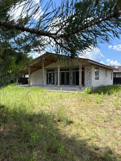 A single-story house under construction is viewed through pine branches in Khmilna, Ukraine. The house is made of cinder blocks and has large glass doors and windows. A covered porch extends across the front of the structure, supported by wooden posts. The surrounding area is overgrown with tall grass and weeds, and the sky above is blue with scattered white clouds.