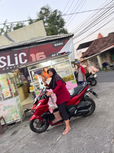 A slightly blurry, medium-shot, eye-level outdoor image captures a woman in a red hijab and long-sleeved red shirt, black pants, and flip-flops. She is positioned on a bright red motorcycle, facing the camera and slightly to the left, with her right hand raised. The motorcycle is parked on a paved surface next to a stall. Behind her, a stall is visible with a large sign displaying "SLIC 16 KRETEK" in white lettering with a red outline. The stall also features a white flag and displays various products and signage. In the background, a man in a grey and red shirt and blue cap is visible near another motorcycle loaded with goods. The scene appears to be a street in Karangtanjung, Indonesia, with buildings and power lines overhead.