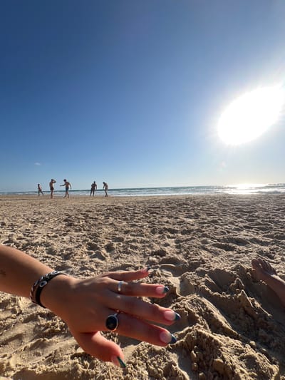 A sunlit beach scene is captured from a low angle. The sky is a bright blue with a prominent sun. The tan-colored sand occupies the right side, where a person's hand with rings and bracelets is visible. Distant figures stand at the water's edge where the sea meets the beach and the sky. The horizon is a slightly crooked line cutting the frame in half.