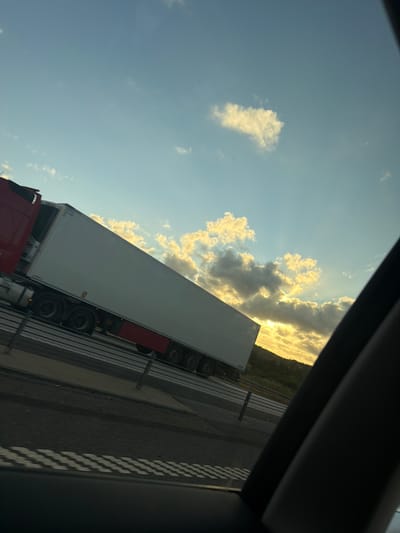 This is an image of a semi-trailer truck on a highway. The truck is red and white and is carrying a white trailer. The truck is moving from left to right. The sky is blue with some clouds. The sun is setting, and the light is coming from the right. The image is taken from inside a car.
The specific location in the city of Uddevalla, Sweden is not identifiable from the image.