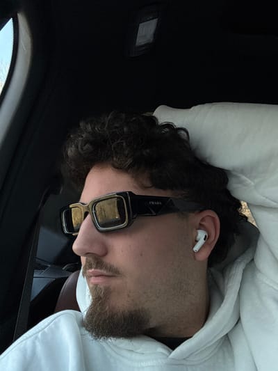 A male individual is depicted in profile, seated in what appears to be the passenger seat of a vehicle in Rahovtsi, Bulgaria. He has dark, curly hair, a mustache, and a short goatee. He is wearing a white hooded sweatshirt, black rectangular Prada brand sunglasses, and a white wireless earbud in his right ear. His head is resting against a white cushion or headrest. The vehicle's dark interior headliner is visible above, along with a rectangular overhead light fixture. A window to the left reveals hints of a natural outdoor environment. A brown interior element is present in the lower background. The individual is looking towards the left side of the frame.
