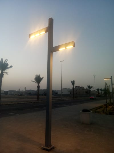 The image shows a dusk or dawn scene in Al Khobar, Saudi Arabia, featuring a modern T-shaped lamppost on a paved walkway. The lamppost emits a warm light. Beyond the walkway is a road with a car driving on it. Palm trees are visible in the background, along with low-rise buildings. A concrete trash receptacle is near the foreground. Other lampposts are visible in the distance. The sky is a pale blue-gray. There are no people visible in the image.
