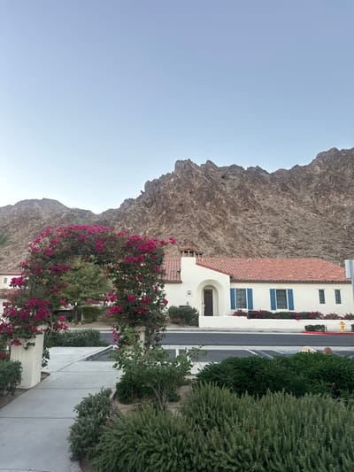 The image displays a clear daylight scene in La Quinta, United States, centered on a building backed by rugged mountains. In the immediate foreground, a concrete archway is heavily covered with vibrant pink bougainvillea flowers, framing a section of a concrete pathway that leads to a paved street or parking area. Various green, low-lying shrubs and bushes are present along the path and street. The main subject is a single-story building constructed with light-colored (likely white or cream) stucco and a terracotta-tiled roof. This building features a prominent arched dark wooden front door, blue-shuttered windows, and a visible house number "77-752" near the entrance. A chimney-like structure extends from the roofline on the left side of the building. A yellow fire hydrant is visible on the far right side of the paved area. The background is dominated by large, rocky, brown and gray mountains with visible geological features, rising sharply under a clear, pale blue sky. No individuals, animals, or vehicles are present or engaged in any actions within the scene.