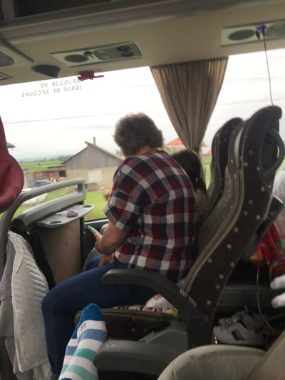 The image depicts the interior of a passenger bus in Fântâna Mare, Romania, viewed from the perspective of a seated passenger. In the immediate foreground, the photographer's feet are visible, clad in white socks with horizontal blue and light blue stripes.

Directly in front, a passenger is seated, their back to the camera. This individual has short, dark, curly hair and wears a short-sleeved plaid shirt with red, white, and dark blue squares, paired with dark blue pants. They are holding a green smartphone in their right hand, looking down at the screen. To the left of this passenger, a grey plastic armrest with an integrated, folded-up tray table is visible. A small, purple, rounded object rests on the tray table surface. A light-colored, possibly quilted garment hangs over the back of the seat to the left.

To the right of the primary seated passenger, another dark brown bus seat is partially visible, featuring a pattern of small light-colored dots and a grey plastic handle at the top. Beneath this seat, a pair of white athletic shoes with dark stripes is discernable on the floor. A multicolored string or ribbon hangs from the seat.

Above the window, a light-colored ceiling panel contains various fixtures, including a speaker or vent grille. A red handle, identified by the French text "DE SECOURS" (Emergency Exit), is affixed to the ceiling. The reflection of this text, "ISSUE DE SECOURS", is visible on the window. Brown, pleated curtains are pulled back from the window on the right side.

The window reveals a rural landscape under an overcast sky. In the middle distance, a traditional-style house or farm building with a gabled brown roof and light-colored walls is prominent, flanked by green fields or pastures. Another, less distinct structure is visible behind it. The outdoor scenery exhibits slight motion blur, indicating the bus is in transit.