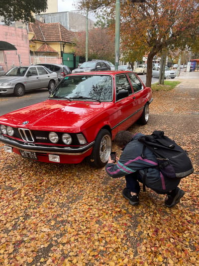 A red BMW 323i automobile is parked on a street covered with fallen leaves, in what appears to be the city of Florida, Argentina. A person wearing a dark jacket with purple accents and a dark backpack is crouched near the front passenger-side tire of the car, holding a small rectangular object, possibly a camera or phone. Several other cars are parked along the street. Buildings, including houses with pitched roofs, line the opposite side of the street. Trees are visible with some leaves remaining on the branches.