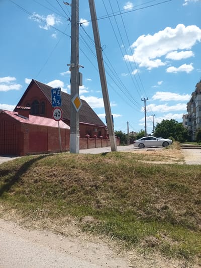 The image shows a street scene in Krymsk, Russia, on a sunny day. In the foreground, dry grass covers a sloped embankment next to a dirt path. A gray concrete utility pole stands prominently on the right, supporting numerous overhead wires and two security cameras. A speed limit sign displaying "40" is attached to the pole, along with a pedestrian and vehicle crossing sign. To the left of the pole, a red brick building with a red tiled roof is visible, featuring arched windows and a smaller attached brick structure. Beyond this building, a white sedan is parked on the side of a paved road. In the background, a multi-story apartment building and lush green trees line the street, under a bright blue sky with scattered white clouds.
