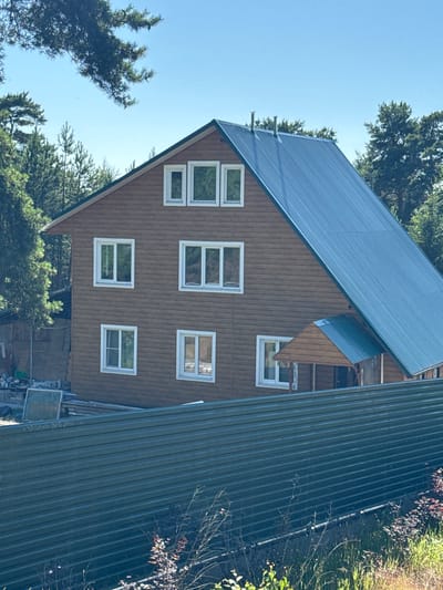 A two-story wooden house with a dark blue corrugated metal roof is visible in the rural settlement of St. Ozerki, Russia. The house has multiple windows with white frames on both visible sides. A covered porch with a similar metal roof is attached to the front right side of the house. In the foreground, a dark green corrugated metal fence runs horizontally across the image, partially obscuring the lower portion of the house and surrounding vegetation. The environment is characterized by a clear blue sky and numerous evergreen trees. Construction materials, including lumber and a blue tarp, are present near the left side of the house, suggesting ongoing work.