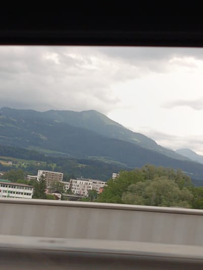 This media file captures a landscape view from what appears to be a vehicle, likely in Hallein, Austria, given the context. The foreground is dominated by a blurred, light gray concrete barrier, suggesting the viewpoint is from a road or highway. Above the barrier, the scene unfolds into a middle ground featuring several multi-story buildings. One prominent building is white with numerous balconies and rows of windows. Adjacent to it are other buildings, some appearing to be hotels or apartment complexes with a similar architectural style of stacked windows. Green trees are interspersed among these structures, and a bridge or elevated roadway is visible in the distance.

The background is characterized by rolling green hills and mountains, covered in dense forests, transitioning to darker, more rugged peaks under a cloudy sky. The overall lighting suggests it is daytime, but the weather is overcast, with a significant amount of cloud cover obscuring the sun. The clouds are a mix of gray and white, creating a diffused light that softens the landscape. The mountains in the distance are shrouded in a hazy, muted blue, indicative of atmospheric perspective and possibly some moisture in the air. There are no visible people or specific activities taking place, presenting a static scene of a semi-urban environment nestled against a natural mountainous backdrop. No discernible text is visible within the image.