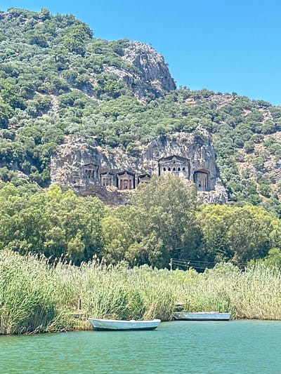 The image shows a scenic view of ancient Lycian rock tombs carved into a cliff face, with lush green trees and vegetation surrounding them. In the foreground, a calm, turquoise body of water stretches along the right side of the frame, with a small part of a boat visible near the shore. Tall reeds and grasses line the water's edge, creating a natural border between the water and the denser foliage further inland. The rock tombs, arranged vertically on the cliff, are a prominent feature, showcasing intricate architectural details despite their age and weathering. The overall scene is bathed in bright sunlight, suggesting a clear and warm day. The presence of the rock tombs indicates a historical or archaeological site, likely a tourist destination given the waterway which could be used for boat tours. The clear blue sky and vibrant greenery contribute to a peaceful and picturesque atmosphere. The location is identified as Ortaca, Turkey, known for its proximity to ancient Lycian ruins. No people or visible text are present in the image.