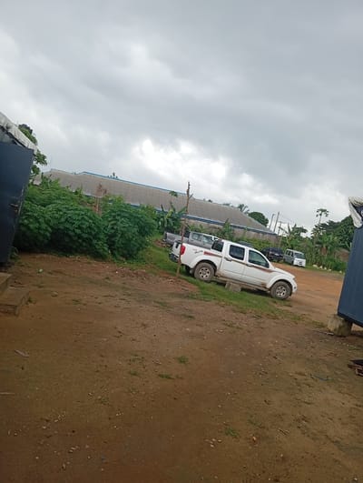 The image depicts an outdoor scene in Igrita, Nigeria. The ground is mostly dirt with sparse patches of grass. In the background, there is a long, low building with a gray roof and green vegetation surrounding it. Several vehicles are parked in the middle ground, including a white pickup truck with red accents. The sky is overcast. There is a dark blue, box-like structure partially visible on both the left and right sides of the frame.