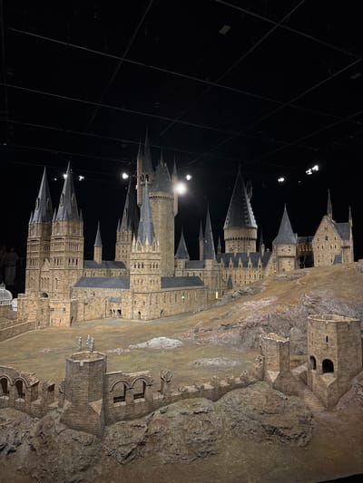 The image shows a detailed model of Hogwarts castle set against a black backdrop with visible lighting rigs. The model is constructed with intricate stonework and features various towers, spires, and walls characteristic of the fictional castle. The model is mounted on a textured base that mimics the rocky terrain surrounding Hogwarts. The scene appears to be a display or exhibit, likely photographed at the Warner Bros. Studio Tour Tokyo, located in Nerima, Japan. The photograph is rotated 90 degrees counter-clockwise.