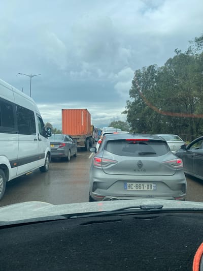 The image depicts a traffic jam on a road in Dar El Beïda, Algeria. The scene is captured from the perspective of someone inside a vehicle, looking out at the traffic ahead. The weather appears overcast. Several vehicles are visible, including a white van, a gray Renault Clio with license plate HC-661-XR 60, a dark gray sedan, and a truck hauling an orange shipping container. The vehicles are closely packed, suggesting heavy congestion. The environment includes trees and a streetlight visible in the background, along with road markings.
