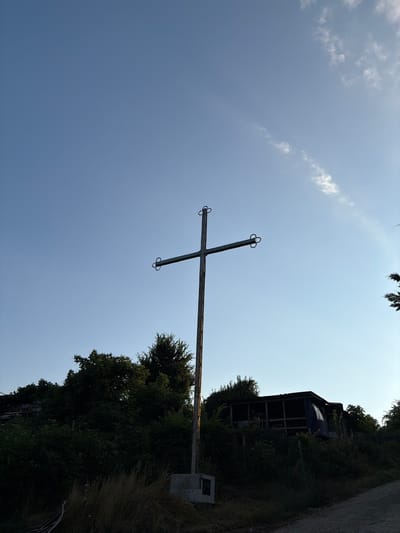 The image depicts an outdoor scene at a low angle, with a clear blue sky dominating the upper portion. In the center, a tall, metallic cross stands prominently, silhouetted against the sky. The cross has a simple design with two curved elements at the ends of the horizontal beam. It is mounted on a concrete base.

In the lower half of the image, dense green foliage and trees form a dark, natural backdrop. Behind the trees, a structure that appears to be a makeshift shelter or building with a wooden frame and a blue tarp is visible. A dirt path or road curves from the lower right corner into the scene. The overall impression is one of a somewhat rural or undeveloped area. The image does not contain any people or visible actions.