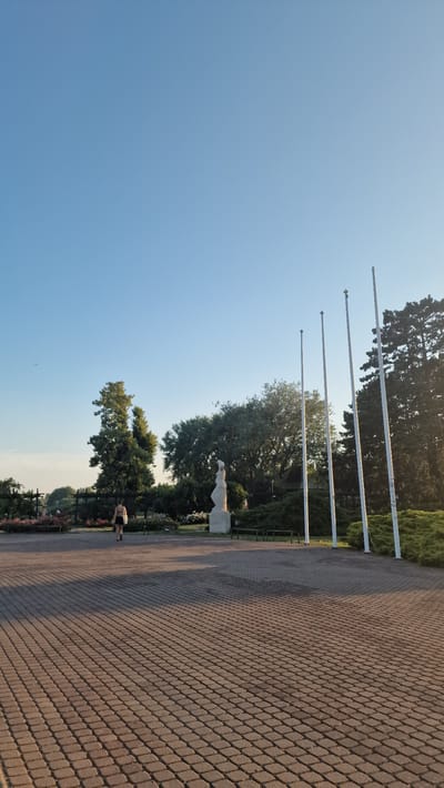 A woman walks across a paved plaza in front of a statue and several flagpoles. The scene is set in a park with trees and flowerbeds in the background. The sky is clear and blue, and the sun casts long shadows across the plaza. This image was taken in Keszthely, Hungary, specifically in the area surrounding the Helikon Palace Museum.