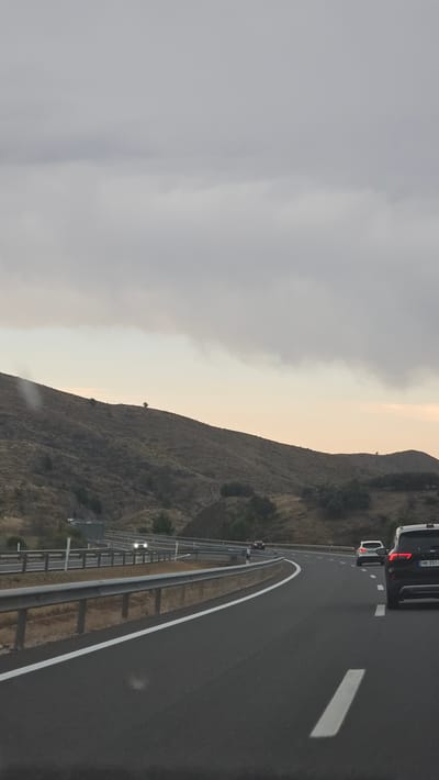 The image depicts a highway scene in Bubierca, Spain, during what appears to be dusk or an overcast day. A grey, cloudy sky dominates the upper portion of the frame. The highway itself is asphalt, with a clear white line marking the lane division on the right side. A silver metal guardrail runs along the left side of the road. In the foreground, a dark-colored SUV is visible, its rear end facing the camera. To the left and further down the road, another white SUV is traveling in the same direction. Further ahead, a car with its headlights on is seen in the distance, approaching on the opposing side of the highway. The landscape flanking the highway is characterized by dry, scrub-covered hills and sparse vegetation. The terrain suggests a rural or semi-arid environment. The overall atmosphere is subdued due to the lighting conditions.