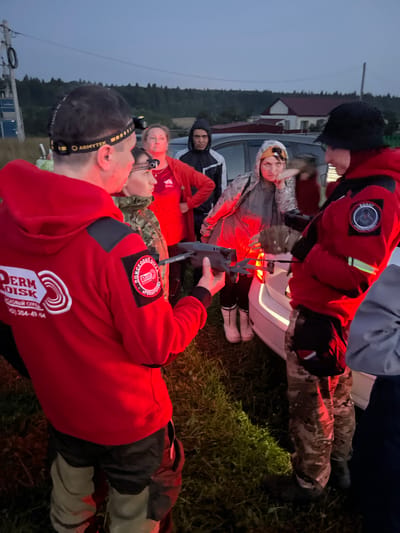 In the evening, in the village of Perebor, Russia, a group of people are gathered outdoors near a white car. One individual in a red jacket holds a drone, seemingly preparing it for flight. Several other individuals, dressed in various outdoor attire including headlamps and camouflage, observe the drone. The background features a rural landscape with trees, a building, and a power line, under a twilight sky. The scene suggests an outdoor activity involving drone operation in a rural setting.