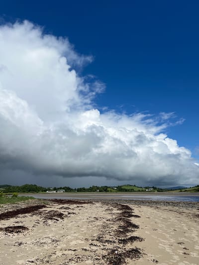 This media file depicts a scenic view of a beach in Dunuisce, Ireland. The image is oriented vertically, showcasing a dramatic sky and a tranquil coastline.
The sky dominates the upper portion of the frame, a vast expanse of deep blue contrasted with voluminous white cumulus clouds. These clouds are so thick and expansive that they appear to stretch across the horizon, with some parts casting a grey, shadowed hue, suggesting a change in weather.
Below the sky lies the sea and the beach. A calm body of water, reflecting the sky's colors, meets a stretch of sandy beach. The sand is a light tan color, interspersed with dark patches of seaweed, likely brought ashore by the tide. The texture of the sand is visible, with subtle footprints or imprints suggesting recent activity.
In the distance, a green, rolling landscape meets the coastline, dotted with scattered white buildings that appear to be houses or small structures, typical of a rural Irish setting. The greenery of the hills suggests a temperate climate. The overall impression is one of natural beauty and serenity, with the dynamic sky adding a touch of drama to the otherwise peaceful scene. The time of day appears to be late morning or early afternoon, judging by the brightness of the sky and the quality of light. The weather is a mix of sun and approaching clouds, creating a visually interesting contrast. There is no visible text in the image.