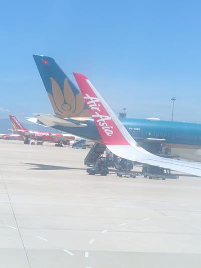 In Bắc Cam Ranh, Vietnam, on a bright, clear day, multiple airplanes are parked on the tarmac. In the foreground, the tail fin of an AirAsia airplane, featuring a red and white design with the "AirAsia" logo, is prominent. Behind it, the tail of a Vietnam Airlines aircraft, adorned with a blue and gold lotus flower motif and a Vietnamese flag, is visible. Several other aircraft, including a red VietJet Air plane with "vietjetair.com" written on its side, are parked in the middle ground. Various airport service vehicles, such as a baggage tug and a mobile stairs unit, are positioned around the airplanes. The scene is set against a backdrop of a light blue sky and a sunlit airport apron.