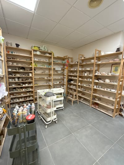 This is a medium-shot, eye-level, wide-angle photograph of a pottery studio, likely located in Mataró, Spain. The room is filled with numerous tall, wooden shelving units, packed with a variety of ceramic pieces in different stages of completion, as well as finished products. The shelves are organized and appear to contain bowls, cups, plates, and decorative items.

In the foreground, a grey, three-tiered rolling cart is visible. It holds a collection of art supplies, including bottles of colorful glazes, brushes in a container, and a red lidded jar. Another similar white rolling cart with three tiers is situated further back in the room, holding various plastic containers and what appear to be ceramic items or molds.

The walls are a neutral, light color, and the ceiling is a white drop ceiling with fluorescent lighting. A circular vent is visible on the ceiling in the upper right portion of the image. The floor is covered in large, grey square tiles with visible grout lines.

The overall impression is one of a functional and organized workspace dedicated to ceramic art. There are no people visible in the photograph.