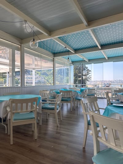This image depicts the interior of an unoccupied restaurant dining area in Marina de Cudeyo, Spain. The space features a structured ceiling with white beams and sections paneled in light blue with a repeating white fan-like pattern. Multiple clear glass orb pendant lights with exposed filaments hang from the ceiling by blue braided cords. The room is enclosed by large, clear glass panels, which extend from a white wooden wainscoting along the lower walls to the ceiling, providing extensive views of the exterior. The floor is dark brown wooden laminate.
Numerous white wooden chairs, each with a light blue seat cushion and a slatted back, are arranged around square and rectangular dining tables. Each table is draped with a white tablecloth topped by a light blue overlay; some tables display white plates. Through the glass panels, a green hillside with white buildings is visible to the left. To the right, a body of water, likely a bay or harbor, is observed, bordered by a distant cityscape featuring various buildings and port infrastructure under a clear blue sky. The scene is illuminated by natural light entering through the glass panels. No individuals are present in the image.