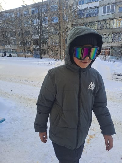 The scene is located in Bolshaya Elhovka, Russia, depicting an outdoor winter environment under daylight conditions. A young individual stands slightly right of center, looking downward and to the left. The individual wears a dark green padded hooded jacket with the hood up over a dark beanie, and large, reflective goggles featuring rainbow-colored lenses covering the eyes. The Kappa logo is visible on the right chest of the jacket. The lower part of the individual's face shows a slight smile. Both hands are bare and hang loosely at their sides. The ground is covered in white snow with visible dirty or melted areas. A light blue object, possibly part of a bench or playground equipment, is present in the lower-left foreground. In the background, a large, multi-story apartment building with numerous windows is visible, partially obscured by bare deciduous trees. Two indistinct figures are visible in the far left background, near a playground structure.