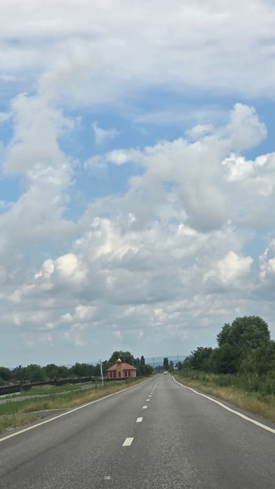 An overcast sky with puffy white clouds is visible above a two-lane asphalt road that stretches into the distance. The road has white dashed lines marking the lanes. On the left side of the road, there is a low, reddish-brown building with a golden dome, possibly a small church or chapel. Beyond the building, there are trees and a wooden structure that appears to be a bridge or embankment. On the right side of the road, dense green trees and vegetation line the route. In the far distance, faint outlines of hills can be seen. The scene is one of a rural or semi-rural road leading through a landscape with a prominent religious building.