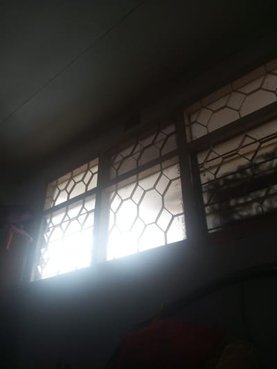 The photograph shows a view from inside a room in Yin Gat Kone, Myanmar, looking up towards three adjacent windows with a decorative lattice frame. Sunlight streams through the windows, creating a bright glare that obscures details of the outside view. The room's ceiling is visible in the upper part of the image. The lower portion of the image is mostly dark, with hints of furniture or other objects.