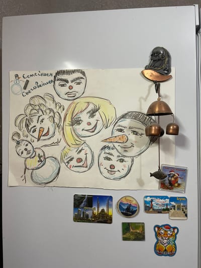 The image displays a collection of magnets and a drawing on a white refrigerator. The drawing, labeled "Семейшен Снеговишек" (Snowman Family), features several stylized snowman faces with various expressions and hairstyles, along with a small, full snowman. Among the magnets, there are several souvenirs from different locations. One magnet shows the city of Grozny, Russia, with a mosque and modern buildings. Another depicts a scene with the text "Счастье и гармонии!" (Happiness and Harmony!) featuring cartoon characters. Other magnets show scenic views and landmarks, including some that appear to be from St. Petersburg and Yalta, Russia. A unique magnet features a metal figurine of a seated figure with a beard, possibly a Buddha or a wise man, attached to a wind chime with small bells and a fish ornament. Another magnet is a small wooden slice with a landscape and the text "Счастье - оно в горах" (Happiness is in the mountains). A small rectangular magnet displays a green landscape. A final magnet features a smiling cartoon tiger wearing a Santa hat and the text "Счастья и радости!" (Happiness and Joy!). The refrigerator is situated against a plain wall. The image is taken from a slightly angled overhead perspective. There is no information about Pokoinoe, Russia, in the image or its contents.