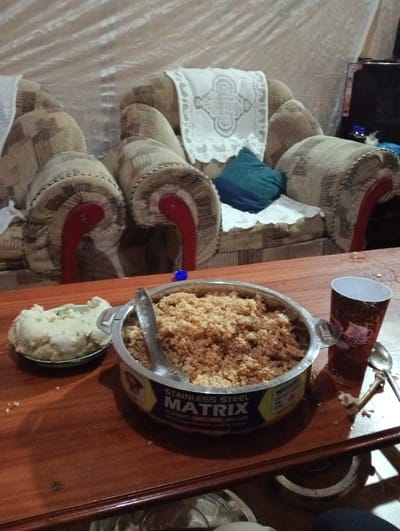 A table setting features a metal container filled with what appears to be couscous, accompanied by a serving of mashed potatoes on a separate plate. Beside the couscous is a disposable cup and a used spoon, indicating a recent meal. In the background, there's a cushioned chair with patterned upholstery, lending a homey atmosphere to the scene.